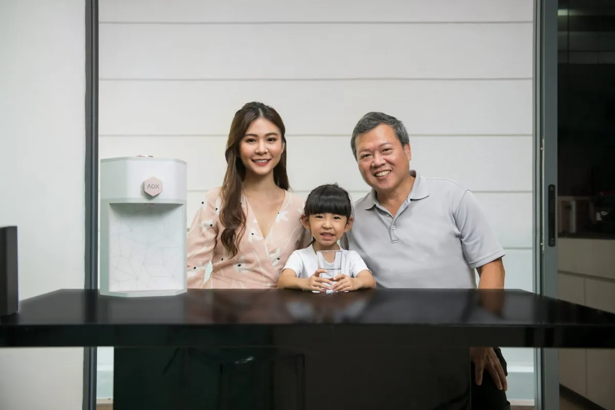 A smiling mother, daughter and grandfather at the table beside their AOX dispenser, the girl holding a glass of water